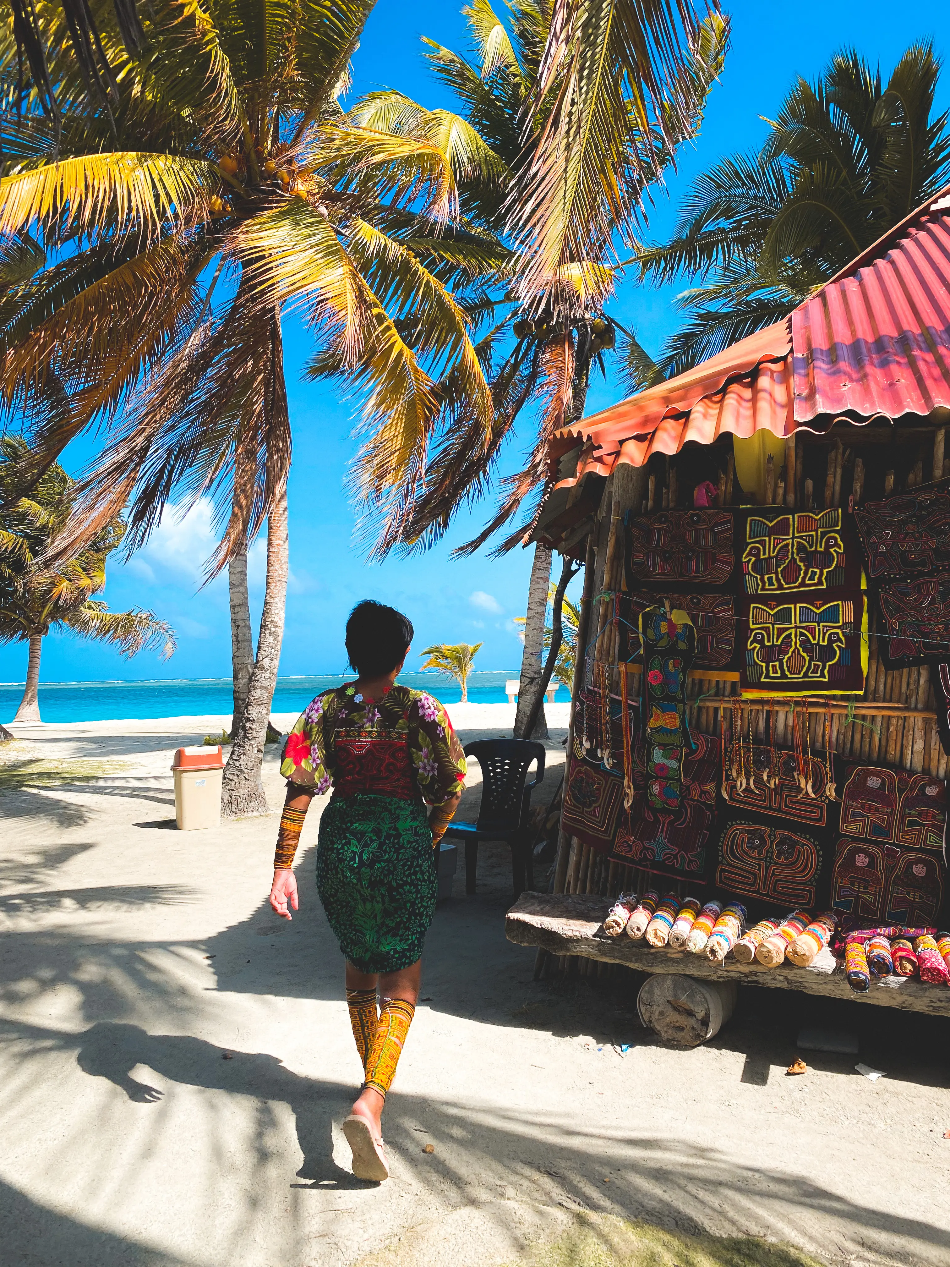 Mujer guna caminando entre palmeras junto a un puesto de molas frente al mar.