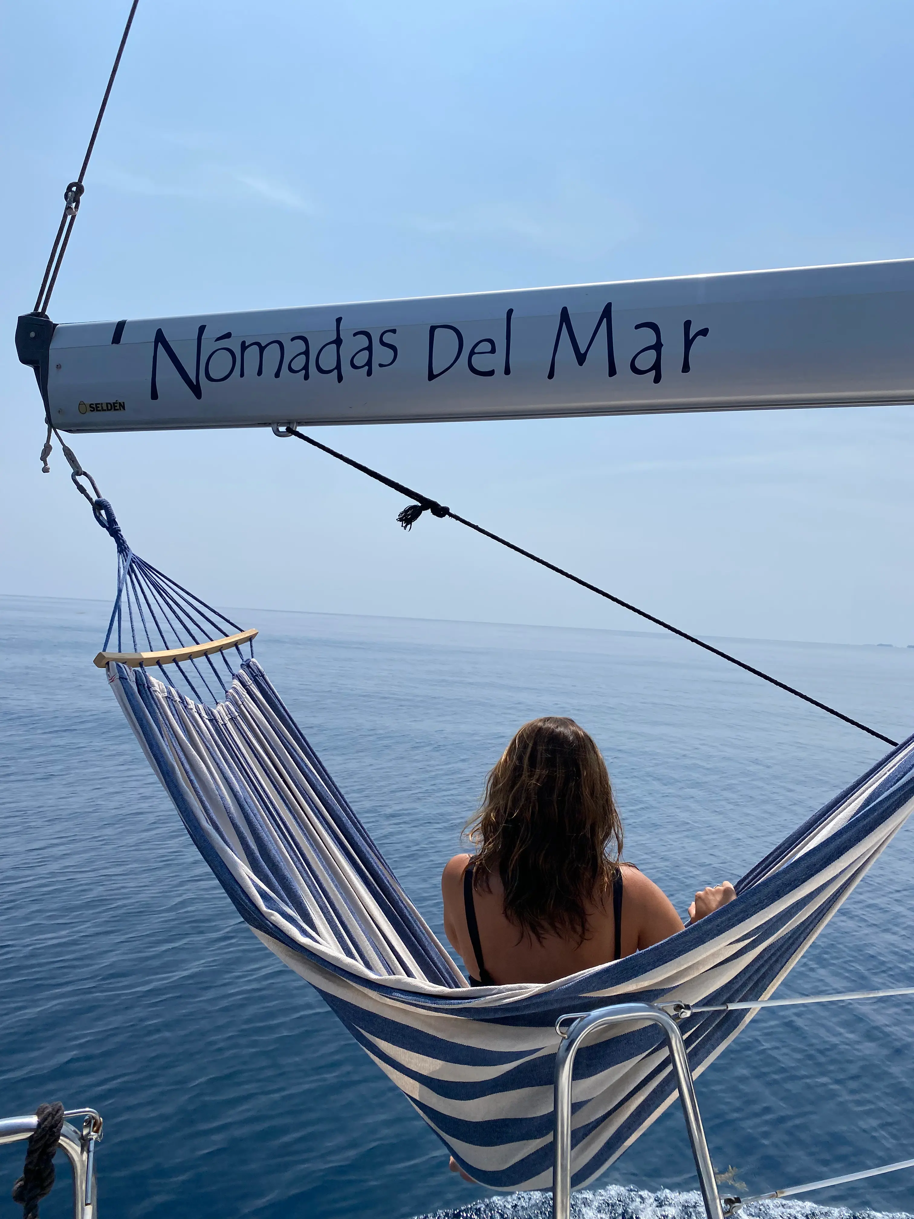 Hombre descansando en una hamaca sobre el agua turquesa junto a un velero.